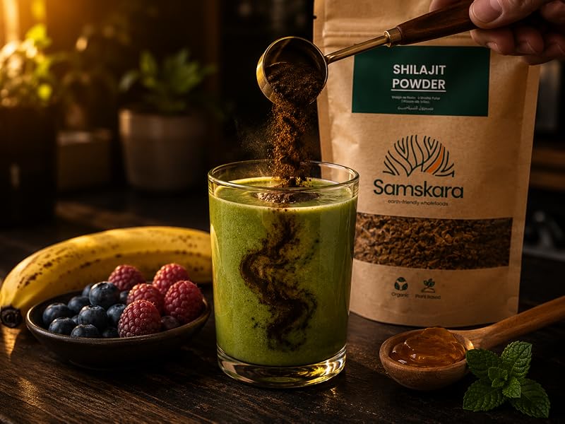Cucharita vertiendo shilajit en polvo en un batido verde con plátano y frutos rojos junto a bolsa Samskara